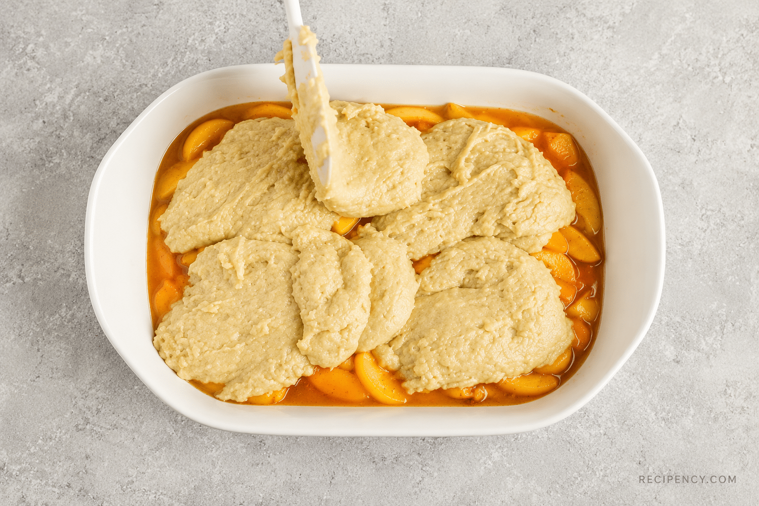 Spoonfuls of cobbler topping placed over spiced peaches in a white baking dish